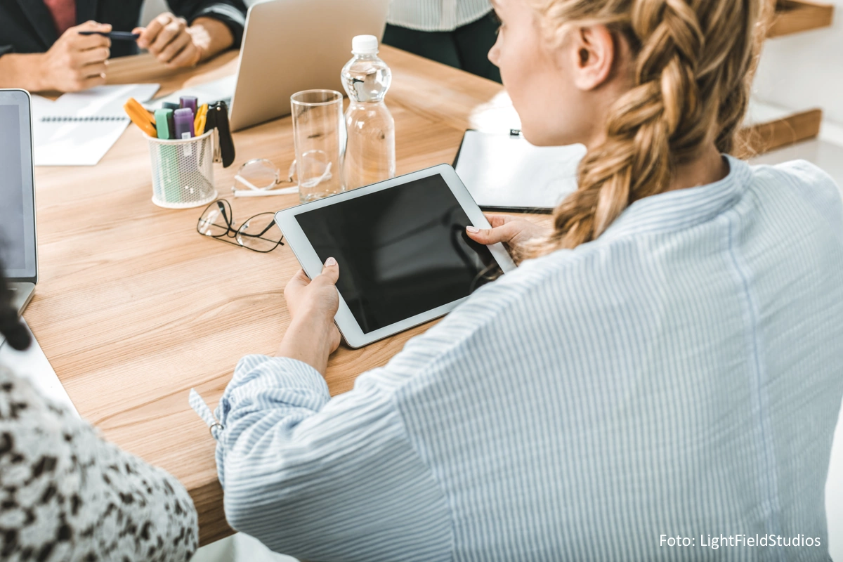 Seitenansicht einer an einem Tisch sitzenden, jungen Frau mit Tablet in der Hand, während eines Meetings
