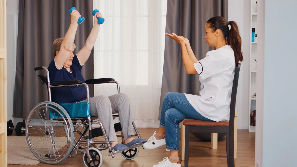 Ältere Frau im Rollstuhl beim Training mit Hanteln während der Rehabilitation mit Krankenschwester. Ausbildung, Sport, Erholung und Aufhebung, Altersheim, Krankenpflege, Gesundheitsfürsorge, Sozialhilfe, Arzt und Heim