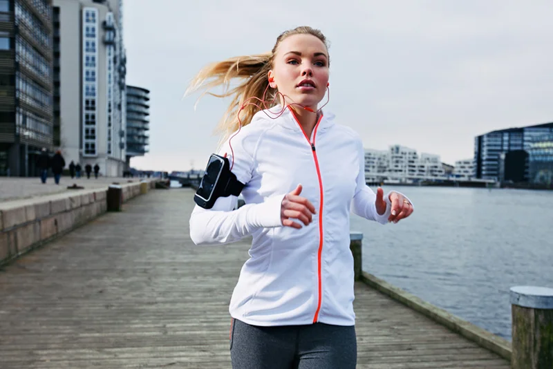 Symbolbild Junge Frau jpggt in der Innenstadt auf der Promenade in Wassernähe und trägt ein Sportarmband um ihr Handy damit zu schützen - Handyhüllen auf Hüllendirekt.de | Stockfoto: envato elements