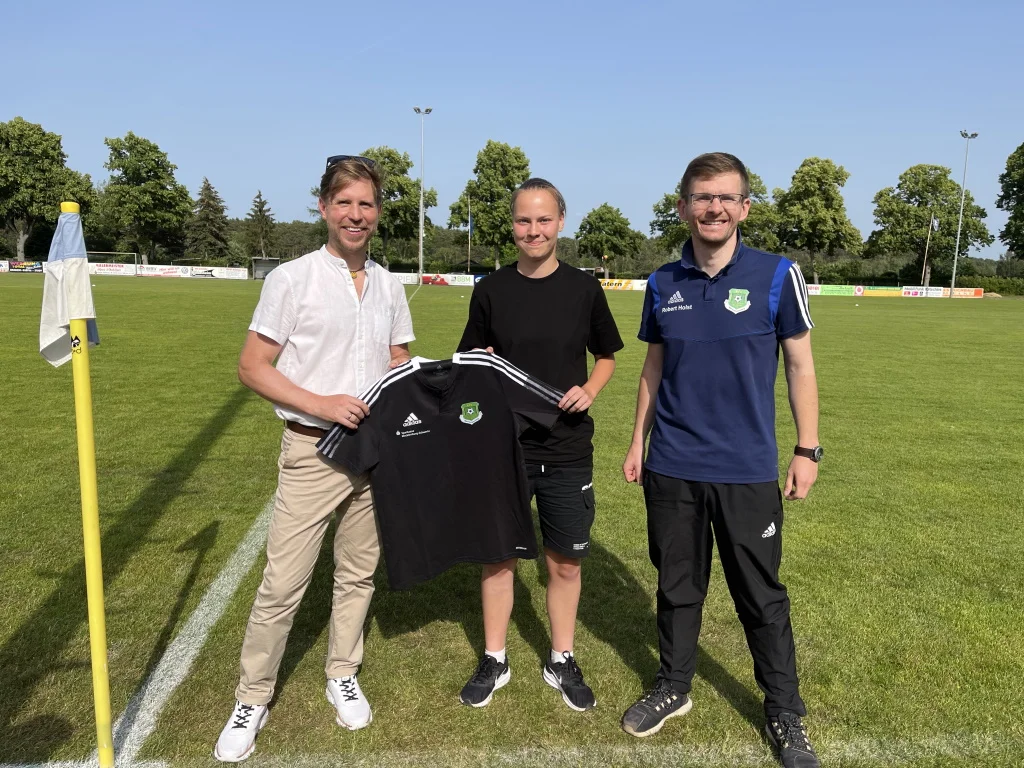 Filialleiter Stefan Pinnow von der Sparkasse Mecklenburg-Schwerin (li.) überreichte die Poloshirts an die Westmecklenburger Schiedsrichter*innen in Person von Hannah Werner von der SG Lindental Tessin und Obmann Robert Holst. (Foto: KFV WM)