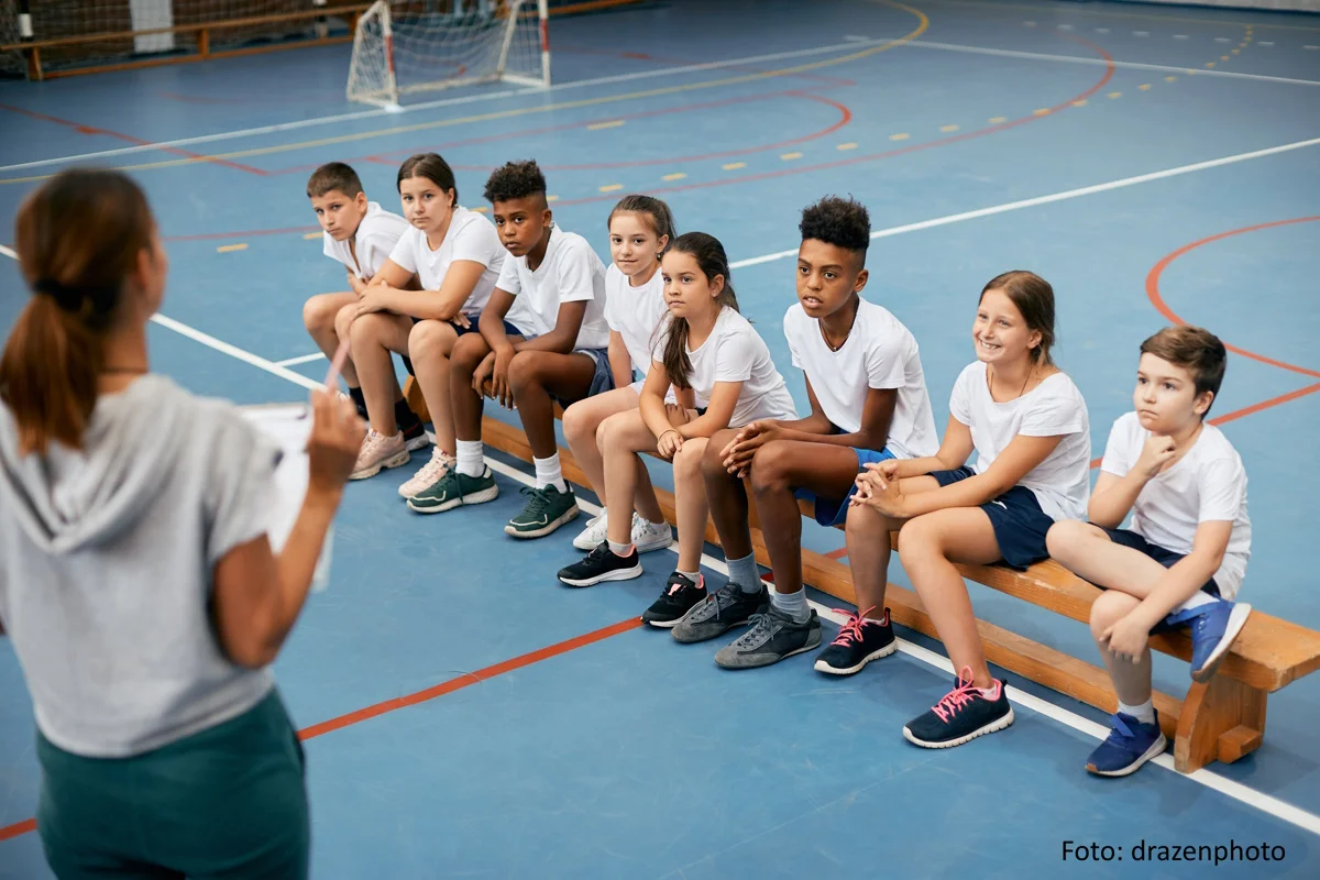 Gruppe von Grundschülern, die Sportunterricht mit Trainerin in der Turnhalle haben.