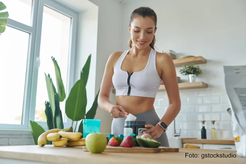 Glückliche junge Frau in Sportkleidung bereitet nach dem Sport sich zu Hause einen Abnehmshake zu und ernährt sich zusätzlich gesund mit Obst, Gemüse