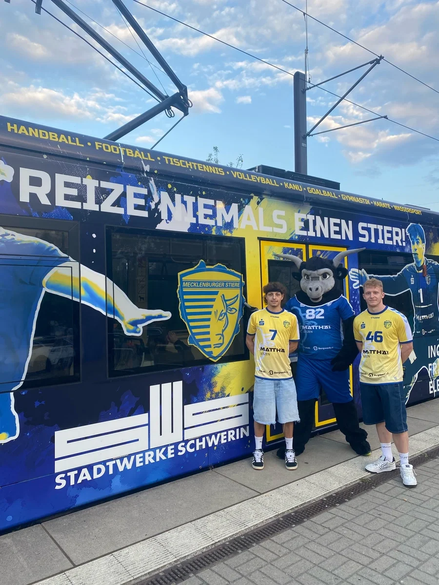Schweriner Handballer und Maskottchen vor der neu im Stiere-Stil beklebten Straßenbahn.
