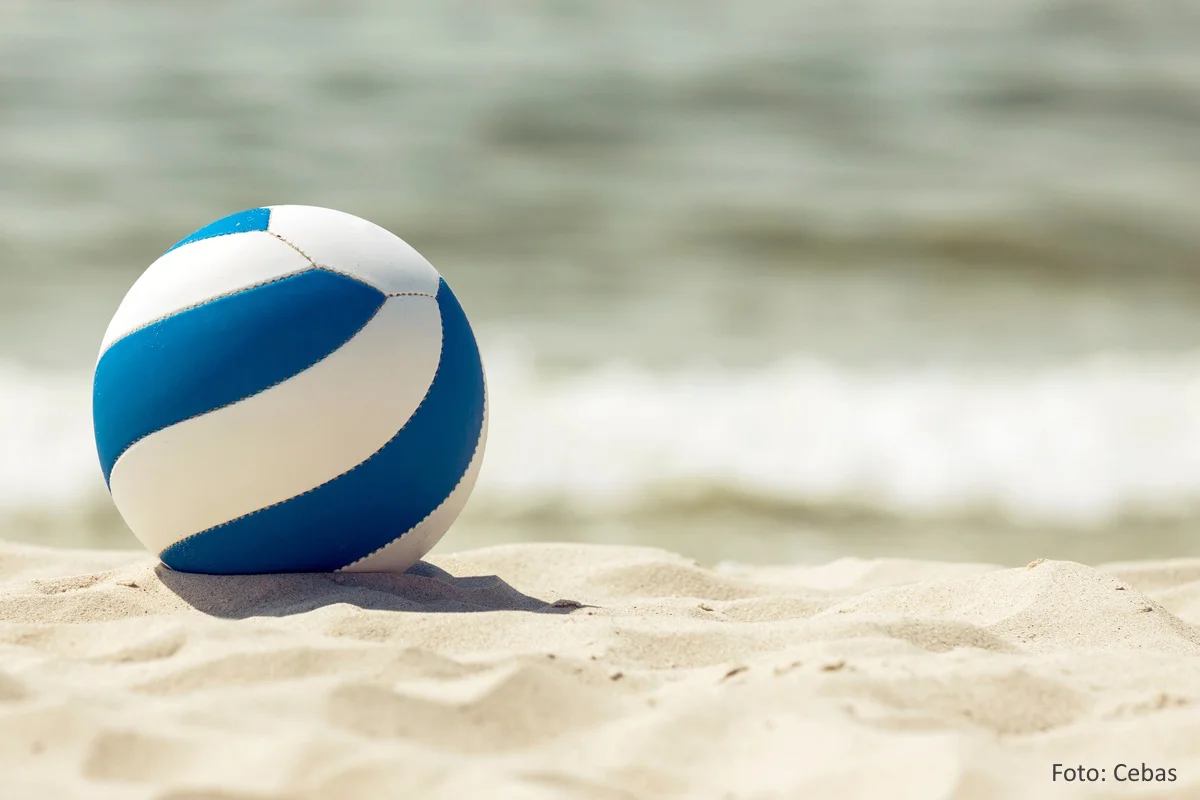 Blau-weißer Beachvolleyball im Sand. Im Hintergrund: das Meer