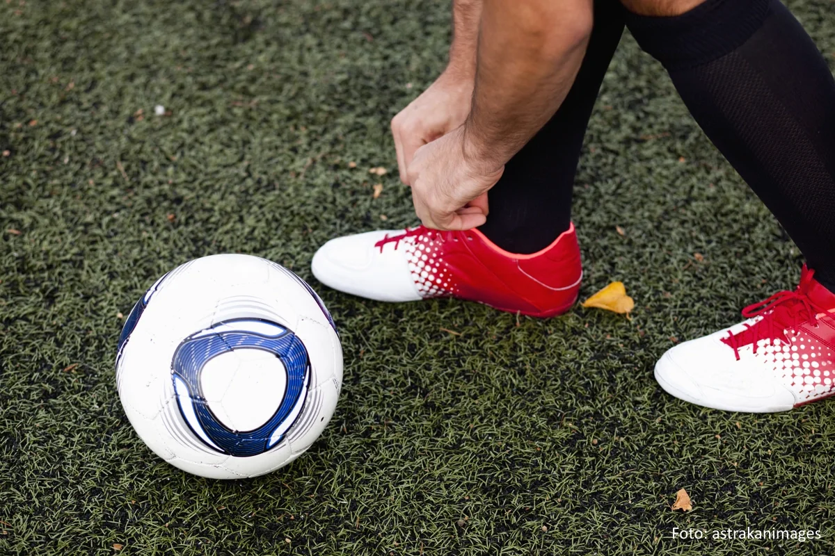 Fußballspieler, der sich auf einem Kunstrasen die Schuhe bindet. Neben ihm liegt ein Fußball.