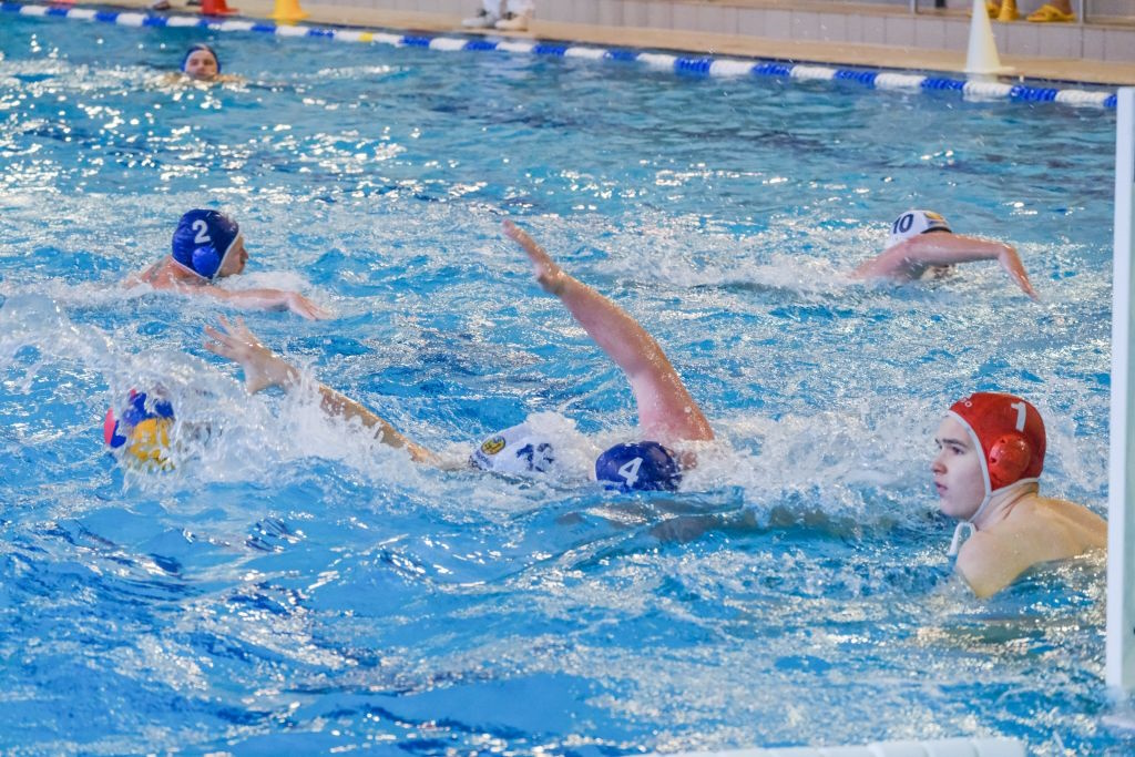 Wasserballer mit blauen und weißen Badekappen schwimmen um den Ballbesitz. Der Torwart trägt rot.