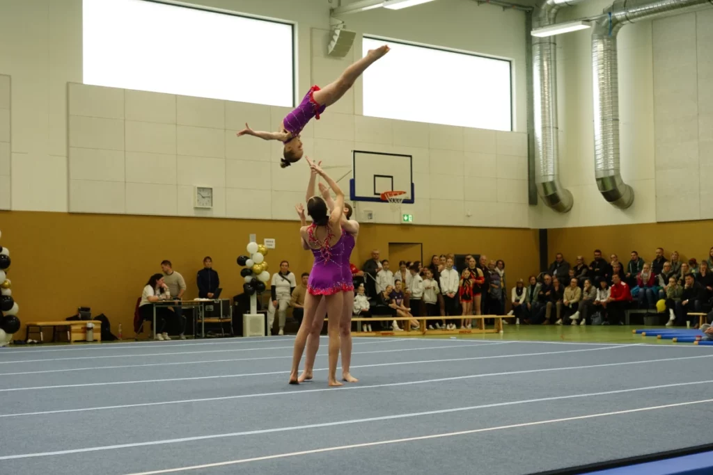 Zwei Schweriner Sportakrobatinnen in Lila Turnanzügen werfen eine dritte Akrobatin gekonnt in die Luft.