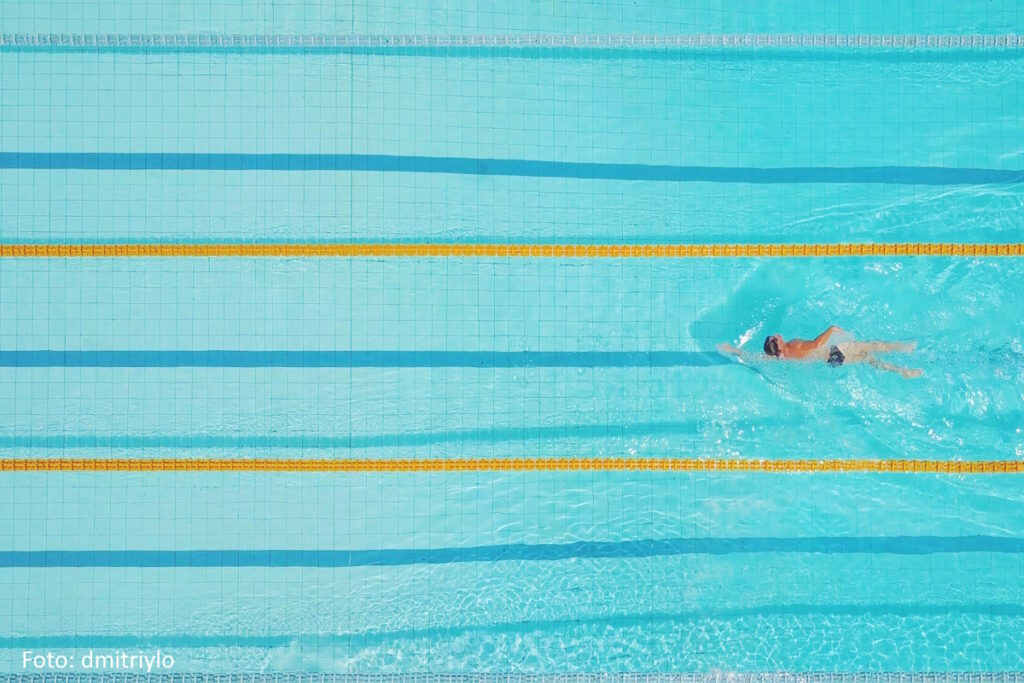 Schwimmbecken von oben. ein einzelner Schwimmer zieht seine Bahnen.