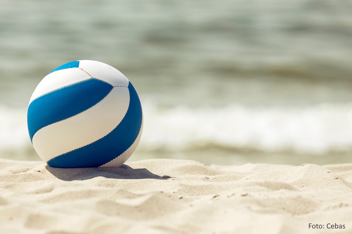 Blau-weißer Beachvolleyball im Sand. Im Hintergrund: das Meer
