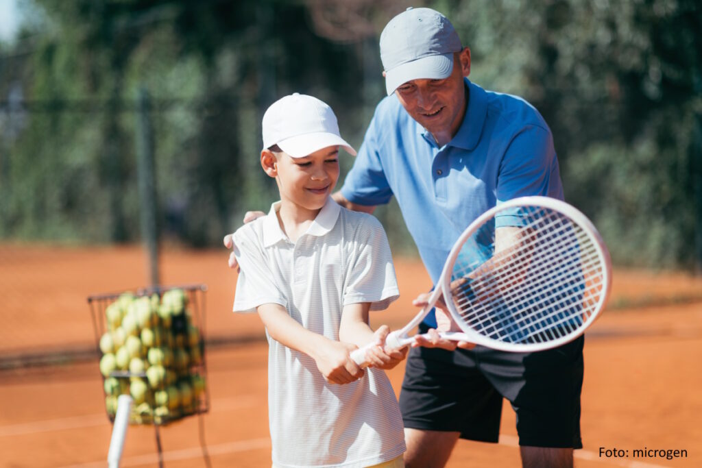 Erfahrener Tennistrainer führt einen Jungen auf dem Sandplatz an den Sport