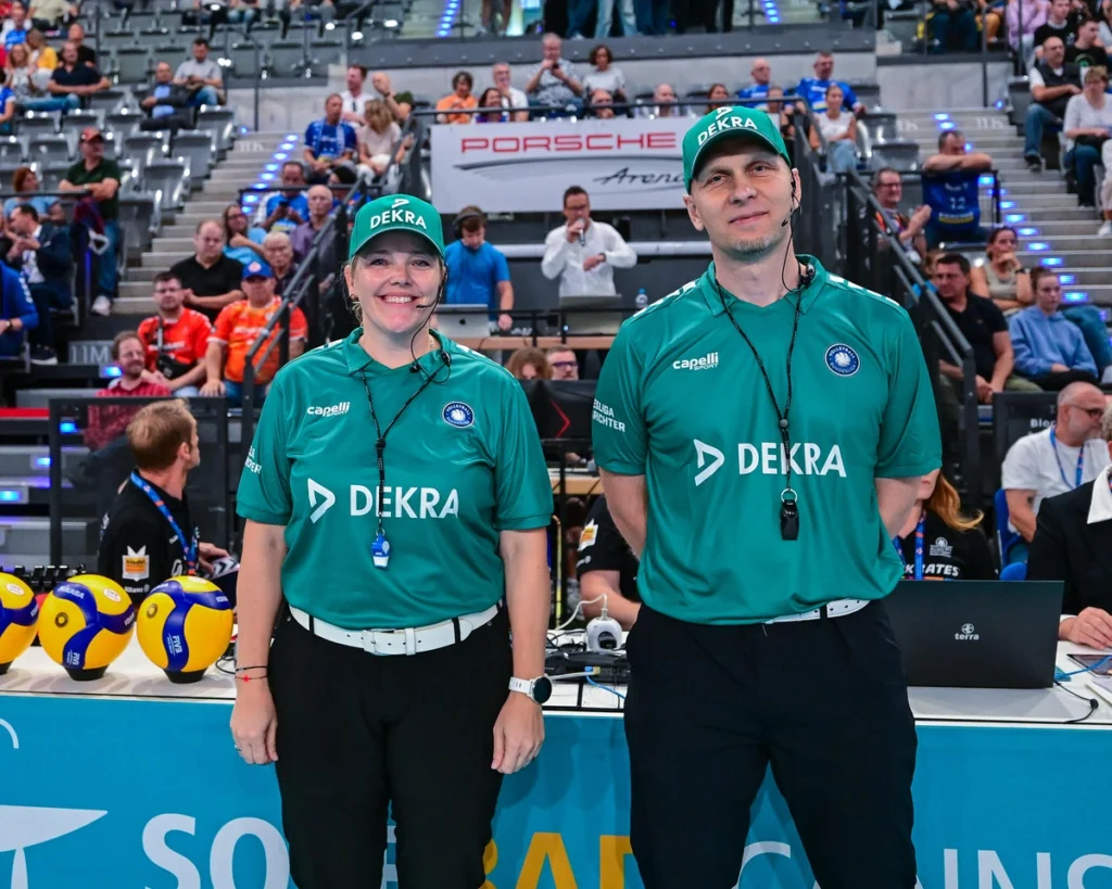 Zwei Volleyball-Schiedsrichterinnen lächeln in die Kamera. Im Hintergrund: die Tribüne mit zahlreichen Zuschauern.