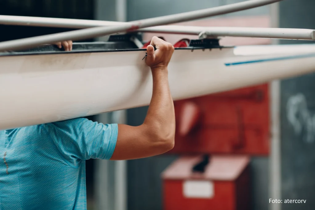 Rudersportler trägt sein Einer-Ruderboot aus der Halle. Zu sehen ist nur der Oberkörper und ein Arm des Sportlers sowei ein Teil seines Bootes.