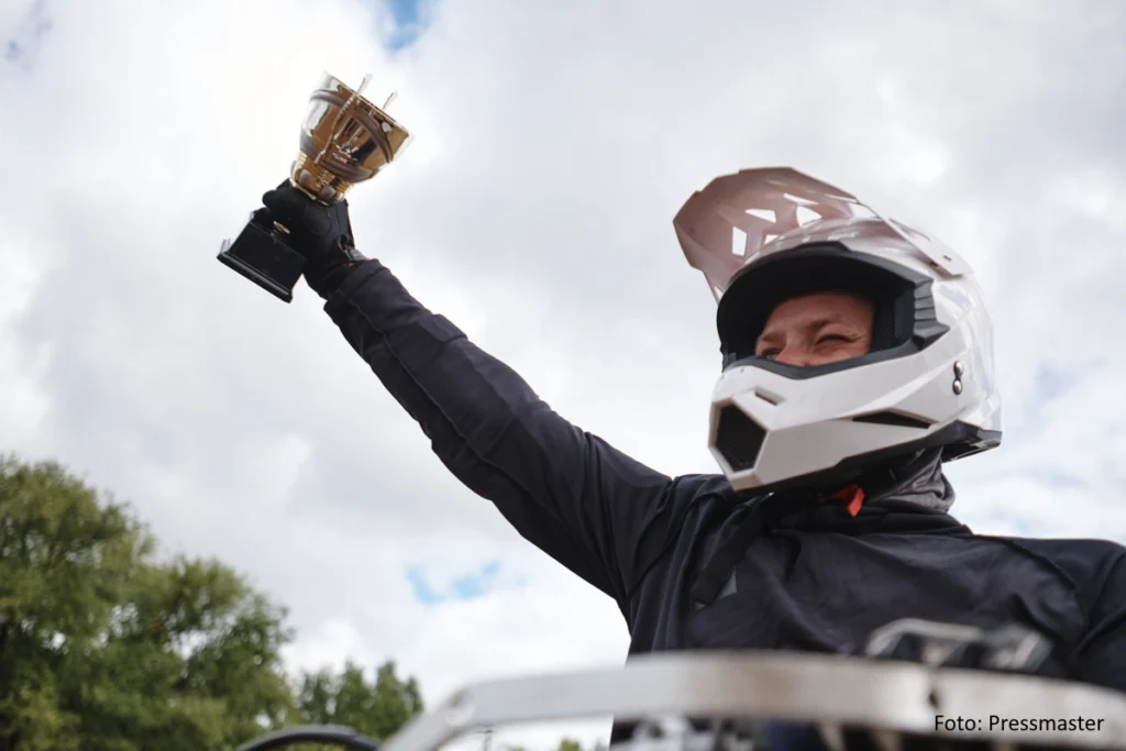 Schwarz gekleideter Motorradfahrer mit weißem Helm reckt einen goldenen Pokal in die Höhe.