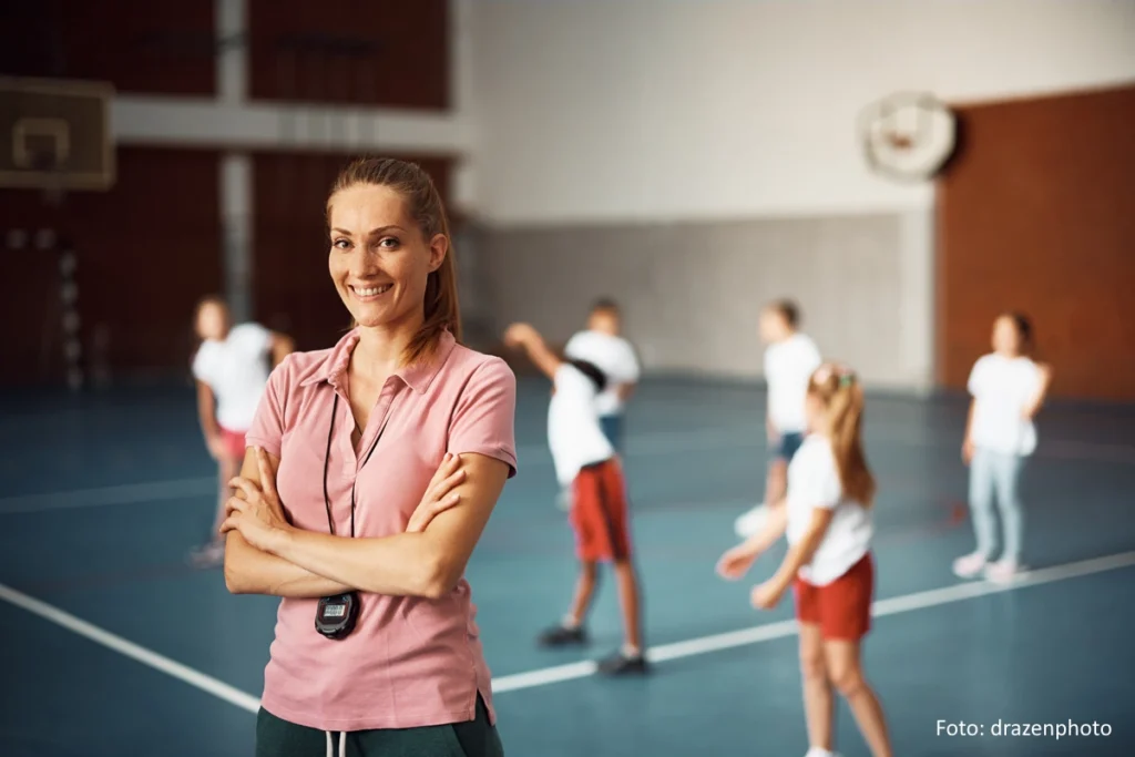 Porträt einer Sportlehrerin, die mit verschränkten Armen in einer Sporthalle steht und in die Kamera schaut. Im Hintergrund erwärmen sich Kinder.